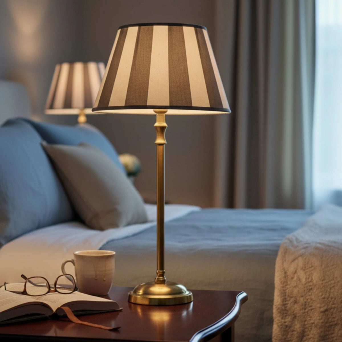 Lampenschirm Livigno in Taupe und Weiß gestreift auf einer Tischlampe mit schlankem goldfarbenem Fuß auf einem Holztisch im Schlafzimmer.