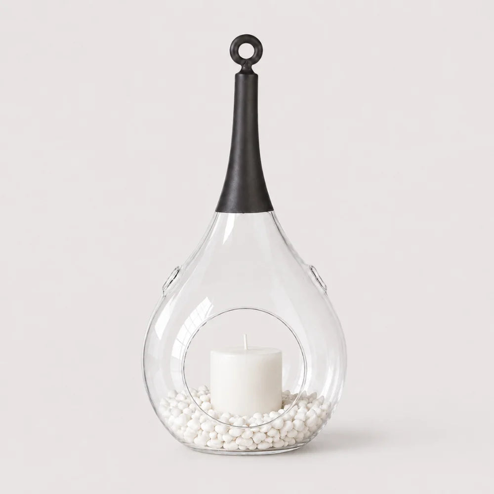 Transparentes Glas-Windlicht mit weißer Kerze, weißen Dekosteinen und schwarzem Griff zum Aufhängen.