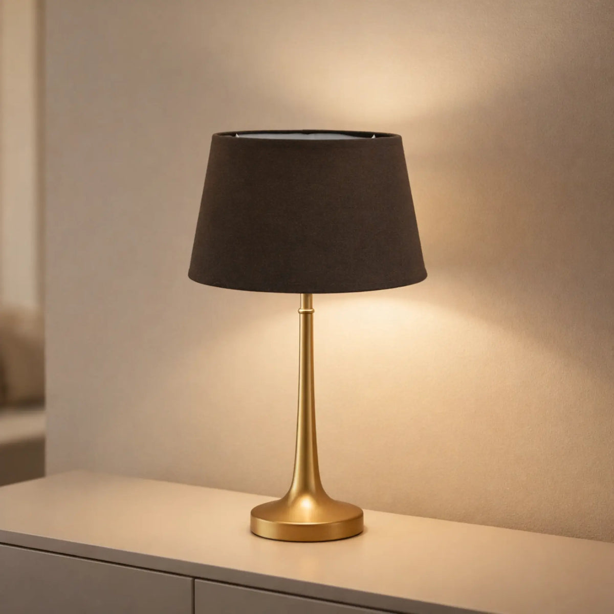 Brauner Lampenschirm aus Baumwolle (Artikel 5825-883) auf einer goldfarbenen Tischleuchte auf einer hellen Kommode, mit warmem Lichtschein an der Wand im Wohnraum.